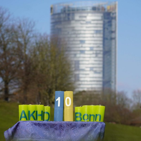 Dieses Foto zeigt unsere Jubiläumskerzen in der Rheinaue mit dem Posttower im Hintergrund. Es sind 10 Kerzen mit jeweils einem Buchstaben oder einer Zahl. Links stehen vier grüne Kerzen mit dem Schriftzug "AKHD", rechts vier weitere grüne mit dem Schriftzug "Bonn", dazwischen zwei hochgestellte mit dem Schriftzug "10". Diese beiden sind in den farben der Ukraine, Blau und gelb, eingefärbt.