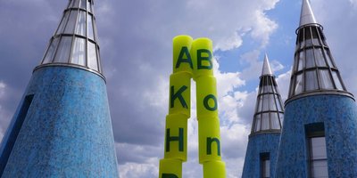 Das Foto zeigt die Jubiläumskerzen als spitzen Kegel aufgestellt. Links stehen die Kerzen mit den Buchstaben A, K, H, D übereinander, rechts die mit dem Schriftzug "Bonn". Unten stehen die Kerzen mit der "10" davor. Im Hintergrund sind die blauen Kegel auf dem Dach der Bundeskunsthalle zu sehen.