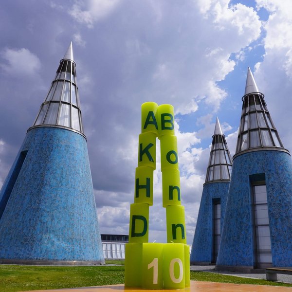 Das Foto zeigt die Jubiläumskerzen als spitzen Kegel aufgestellt. Links stehen die Kerzen mit den Buchstaben A, K, H, D übereinander, rechts die mit dem Schriftzug "Bonn". Unten stehen die Kerzen mit der "10" davor. Im Hintergrund sind die blauen Kegel auf dem Dach der Bundeskunsthalle zu sehen.