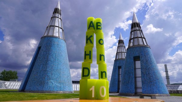 Das Foto zeigt die Jubiläumskerzen als spitzen Kegel aufgestellt. Links stehen die Kerzen mit den Buchstaben A, K, H, D übereinander, rechts die mit dem Schriftzug "Bonn". Unten stehen die Kerzen mit der "10" davor. Im Hintergrund sind die blauen Kegel auf dem Dach der Bundeskunsthalle zu sehen.