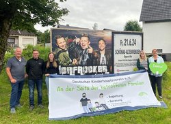 Foto einer Gruppe Menschen auf einer grünen Wiese vor einer Plakatwand. Auf der Plakatwand das Foto der Musikgruppe Dorfrocker. Links im Bild zwei Männer und eine Frau, rechts im Bild zwei Frauen, zwischen ihnen ist eine Bannerplane gespannt. Darauf der Text: Deutscher Kinderhospizverein e.V. - An der Seite der betroffenen Familien.