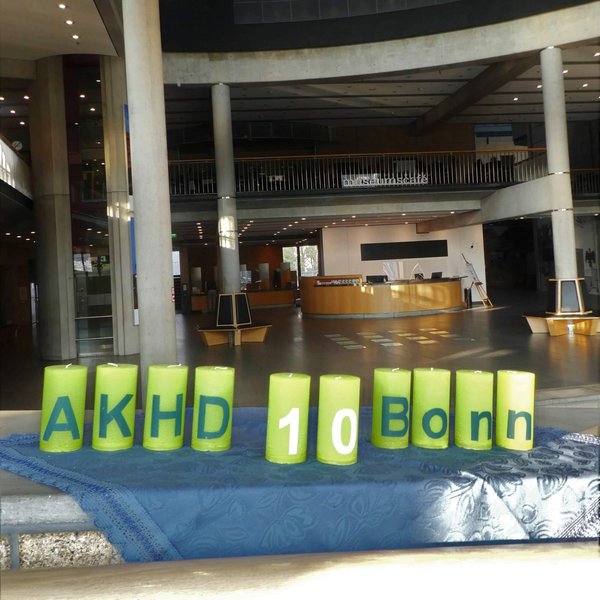 Hier stehen die grünen Jubikäumskerzen mit der Aufschrift "AKHD 10 Bonn" (die "10" steht in der Mitte) auf einer blauen Decke auf einer Mauer im Haus der Geschichte; im Hintergrund das Museumscafé